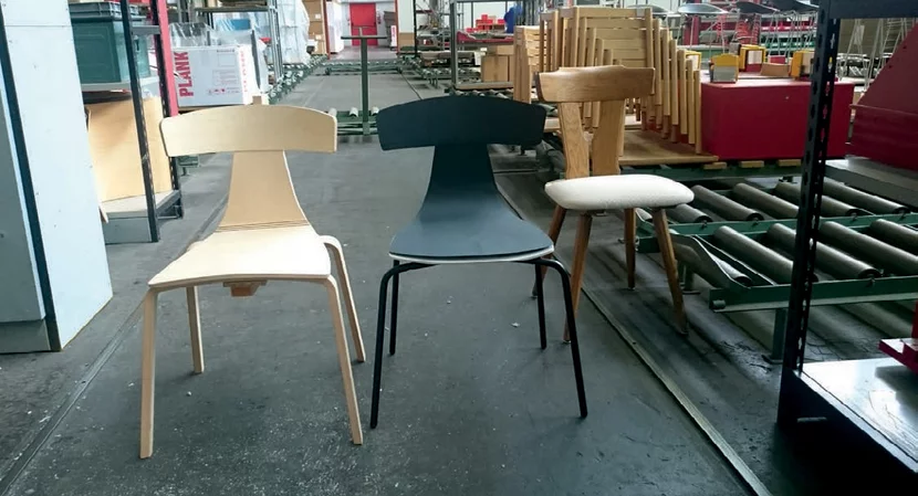 Ein REMO aus Formholz natur und ein REMO mit schwarzer Schale auf Metallbeinen neben einem Holzstuhl in einer Fabrikhalle