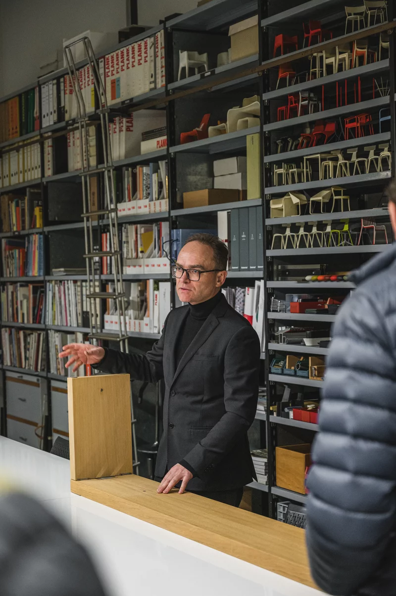 Michael Plank in the PLANK library, in front of shelves with chair models and catalogues, presenting a piece of solid wood